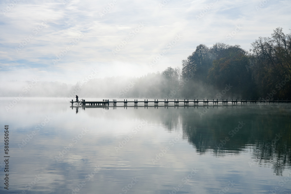 Jetty at the lake in the fog. Misty landscape in the morning. Idyllic nature by the water. Rest and relaxation.
