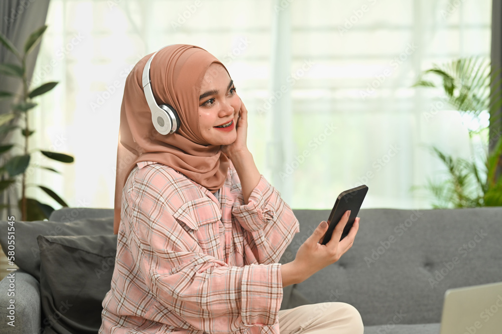 Attractive young woman in hijab holding smartphone and listening to music on headphone, spending free time in living room Attractive young woman in hijab holding smartphone and listening to music on headphone, spending free time in living room