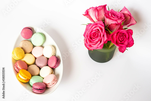 Colorful macaroons on a white ceramic plate and pink roses in a green vase on a white background.