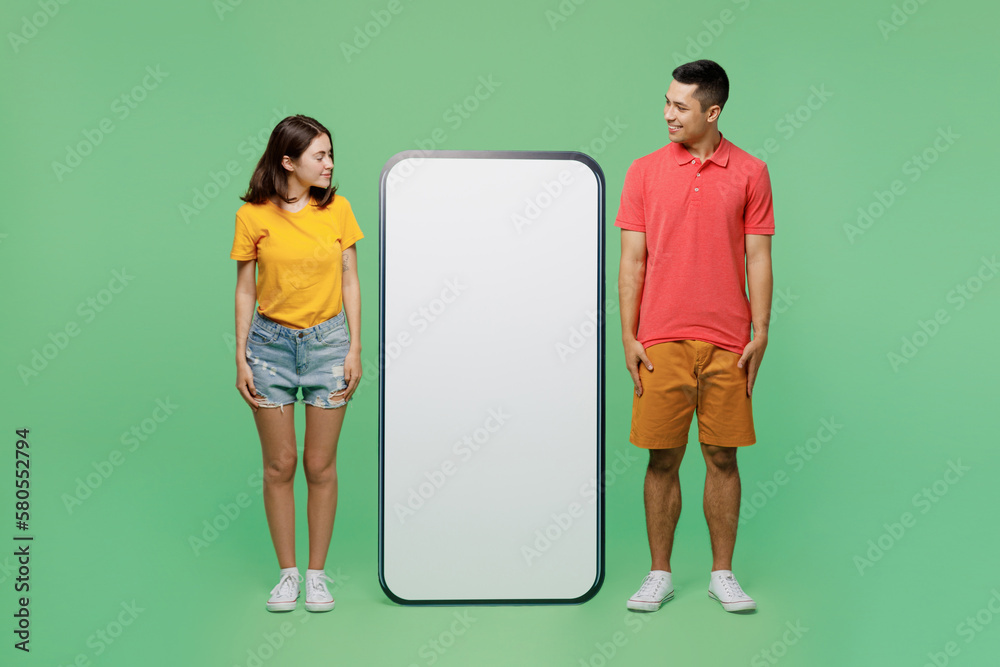 Full body smiling happy fun young couple two friends family man woman wear t-shirts together look at big huge blank screen mobile cell phone with area isolated on pastel plain light green background. Full body smiling happy fun young couple two friends family man woman wear t-shirts together look at big huge blank screen mobile cell phone with area isolated on pastel plain light green background.