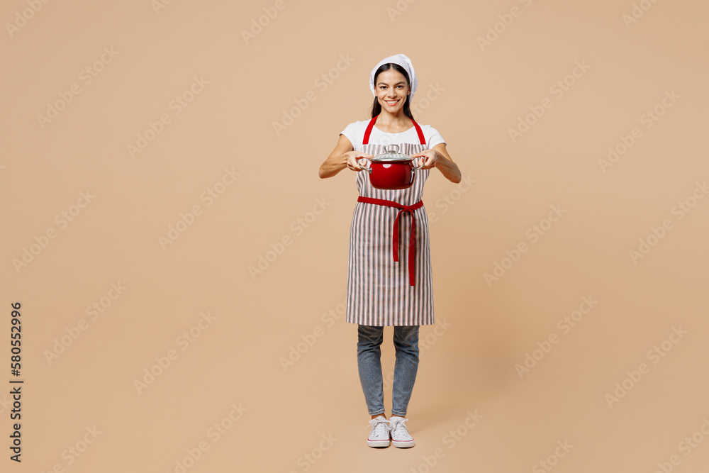 Fototapeta premium Full body smiling happy fun young housewife housekeeper chef baker latin woman wear apron toque hat hold in hand pot pan look camera isolated on plain pastel light beige background. Cook food concept.