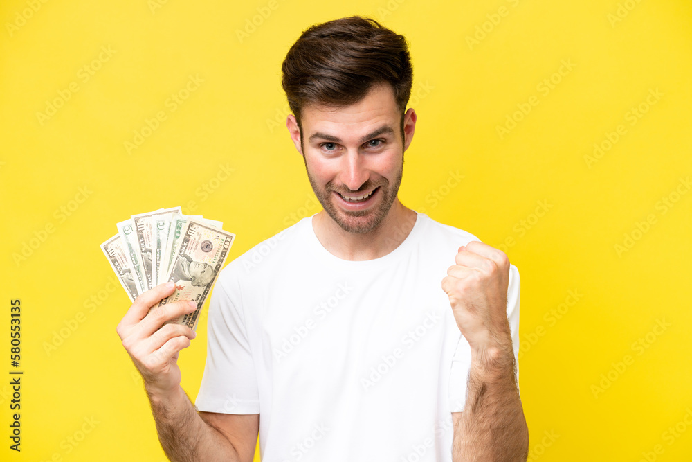 Young caucasian man taking a lot of money isolated on yellow background celebrating a victory in winner position Young caucasian man taking a lot of money isolated on yellow background celebrating a victory in winner position