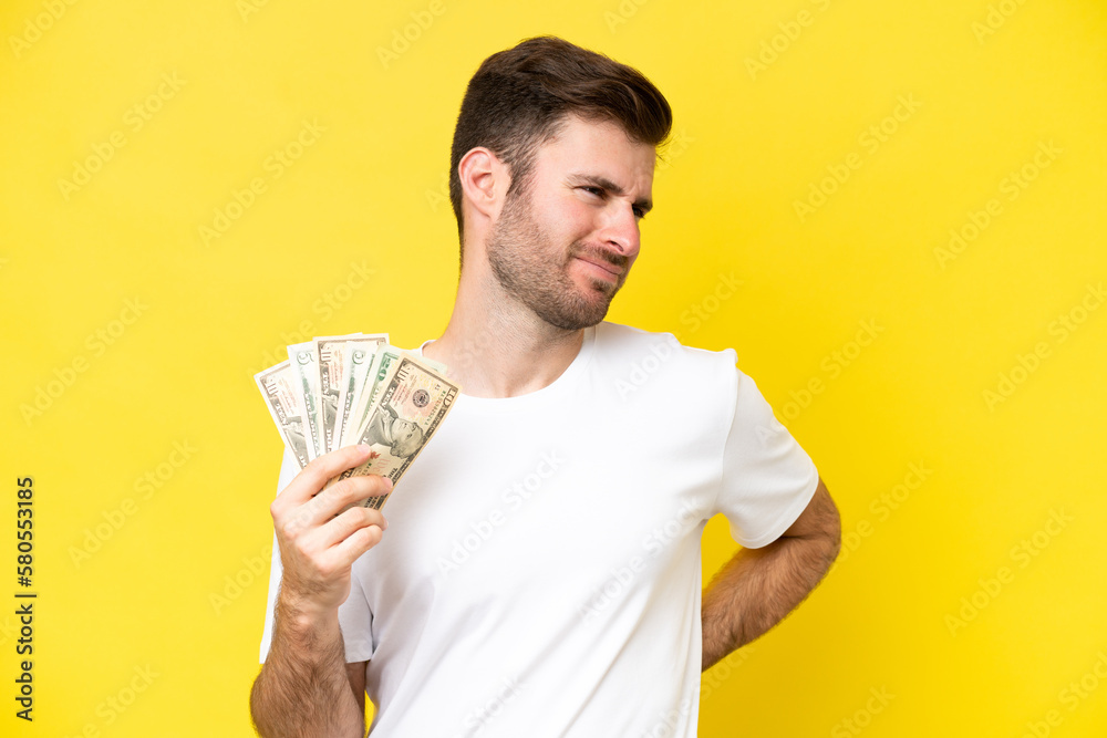 Young caucasian man taking a lot of money isolated on yellow background suffering from backache for having made an effort Young caucasian man taking a lot of money isolated on yellow background suffering from backache for having made an effort
