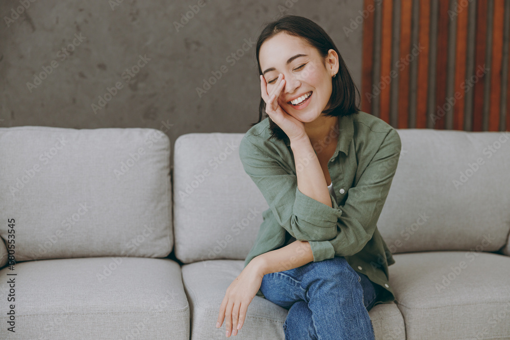 Young smiling shy happy woman of Asian ethnicity wears casual clothes look camera prop up chin sits on grey sofa couch stay at home hotel flat rest relax spend free spare time in living room indoor. Young smiling shy happy woman of Asian ethnicity wears casual clothes look camera prop up chin sits on grey sofa couch stay at home hotel flat rest relax spend free spare time in living room indoor.