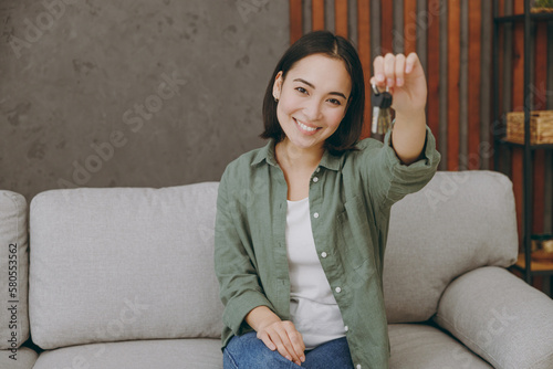 Young fun woman of Asian ethnicity wears casual clothes hold in hand bunch of keys look camera sits on grey sofa couch stay at home hotel flat rest relax spend free spare time in living room indoor.