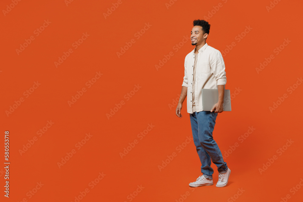 Naklejka premium Full body side profile view student young IT man of African American ethnicity wear light shirt casual clothes hold closed laptop pc computer walk go isolated on orange red background studio portrait.