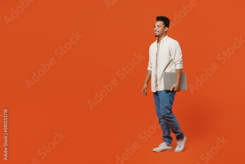 Full body side profile view student young IT man of African American ethnicity wear light shirt casual clothes hold closed laptop pc computer walk go isolated on orange red background studio portrait.