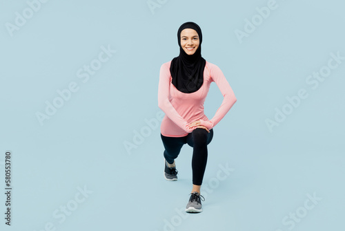 Full body happy young asian muslim fitness trainer sporty woman wear pink abaya hijab spend time in gym look camera do squates isolated on plain blue background studio. Workout sport fit abs concept.