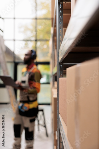 Selective focus of metallic shelves full with clients orders putting in cardboard boxes. African american manager wearing headphones listening music while working at merchandise logistics