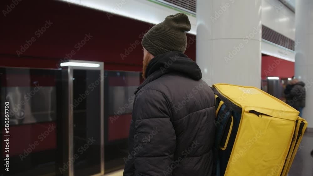 A view from the back of a delivery man with a yellow backpack who is waiting for a subway car A view from the back of a delivery man with a yellow backpack who is waiting for a subway car