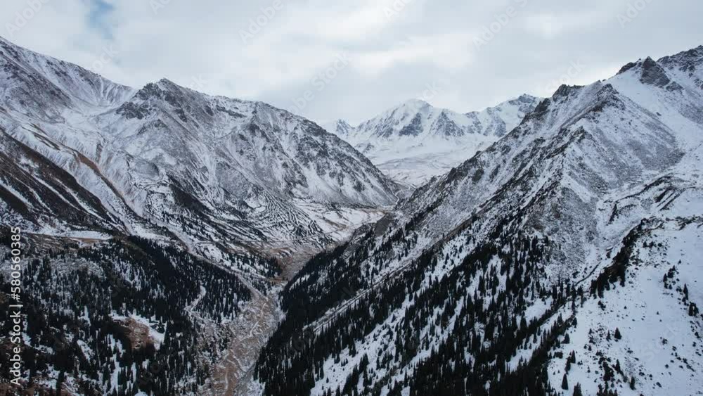High snowy mountains with forest in the gorge. Dark sky with clouds, gloomy atmosphere. The white slopes are covered with snow, high-altitude spruce trees grow. Trails are visible. Big Almaty Lake High snowy mountains with forest in the gorge. Dark sky with clouds, gloomy atmosphere. The white slopes are covered with snow, high-altitude spruce trees grow. Trails are visible. Big Almaty Lake