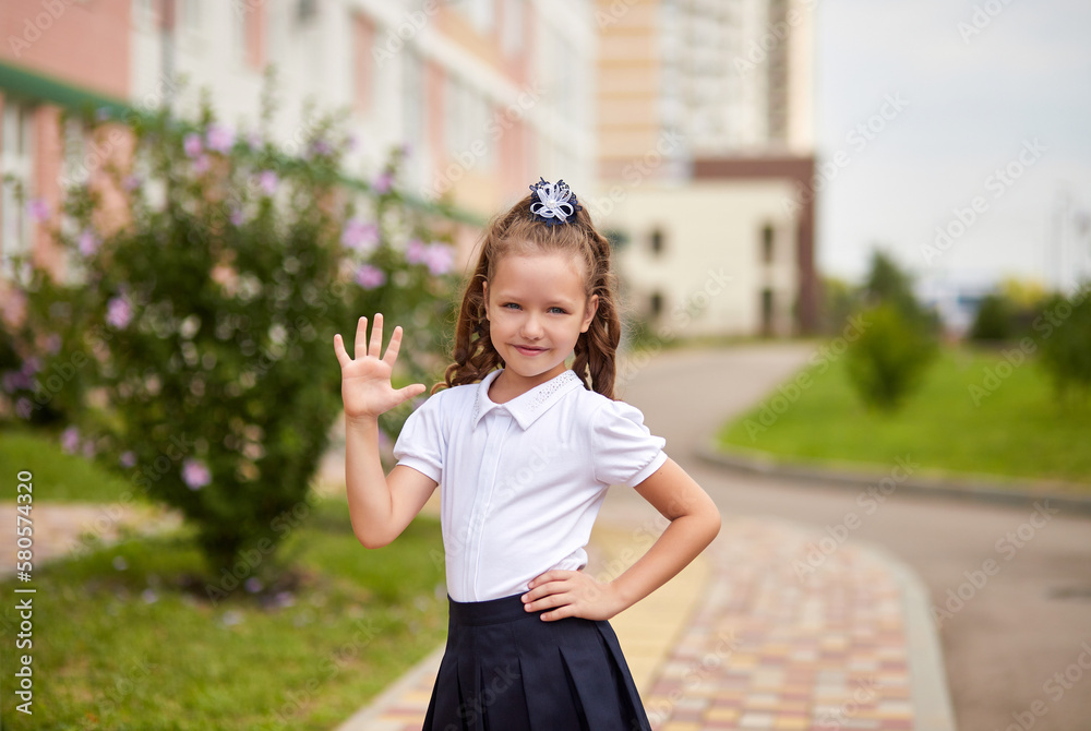 Lovely smiling first grader girl in school uniform shows hi five gesture near the school building. Happy little schoolgirl is ready to go to class for the first time. Lovely smiling first grader girl in school uniform shows hi five gesture near the school building. Happy little schoolgirl is ready to go to class for the first time.