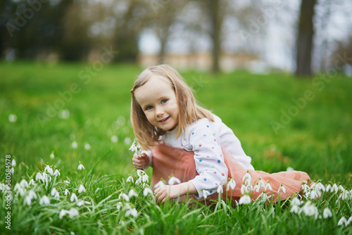 Preschooler girl in pink tutu skirt sitting in the grass with many snowdrop flowers in park or forest on a spring day