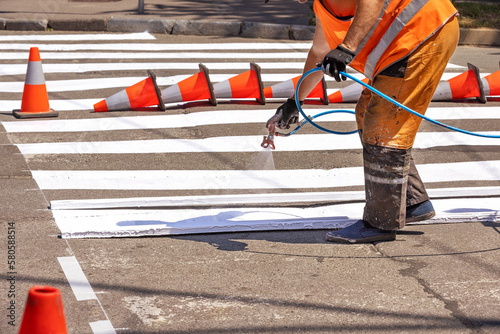 A road worker paints pedestrian markings on an old asphalt road on a summer day.