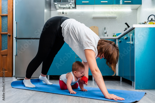 Home yoga. A young Caucasian mother does Adho Mukha Shvanasana while her baby crawls under her on the mat. The concept of sports activity with kids
