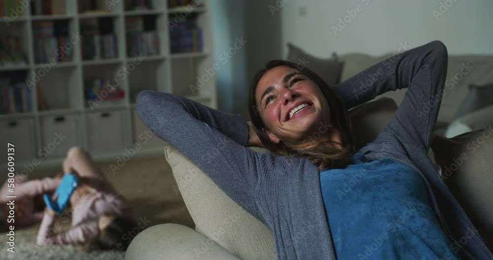 Portrait of a Woman Relaxing on a Sofa at Home While her Two Children are Playing with a Tablet in the Background. Young Mother Enjoying her Weekend, Resting while her Daughters are Busy Playing Games Portrait of a Woman Relaxing on a Sofa at Home While her Two Children are Playing with a Tablet in the Background. Young Mother Enjoying her Weekend, Resting while her Daughters are Busy Playing Games