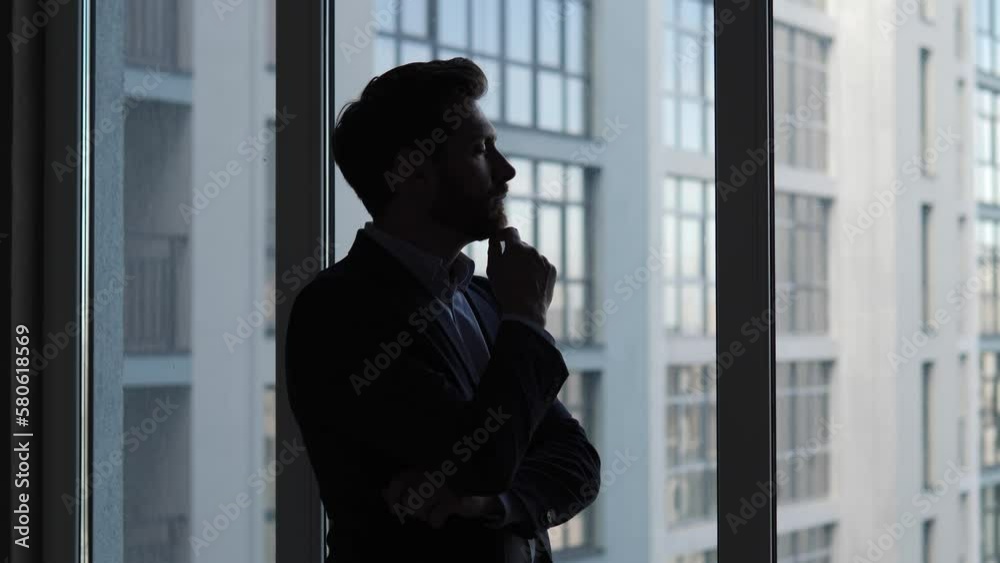 Silhouette of a businessman standing by the window in a skyscraper, lost in thought. Man is contemplating dreaming about his next big move in the business world. Concepts of planning, strategy, ideas. Silhouette of a businessman standing by the window in a skyscraper, lost in thought. Man is contemplating dreaming about his next big move in the business world. Concepts of planning, strategy, ideas.