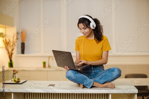 Close up portrait of indian or arabian woman inside home with headset for video call, woman smiling and looking at laptop, couch customer support tech helpline.