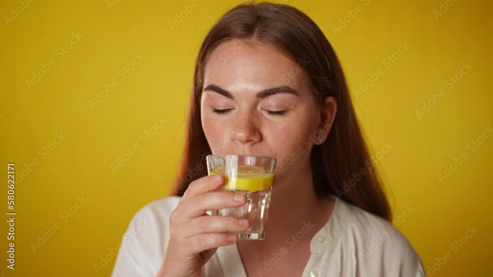 Young woman drinking lemon water smiling looking at camera in slow motion. Portrait of brunette confident Caucasian lady with gray eyes posing at yellow background with healthful drink Young woman drinking lemon water smiling looking at camera in slow motion. Portrait of brunette confident Caucasian lady with gray eyes posing at yellow background with healthful drink