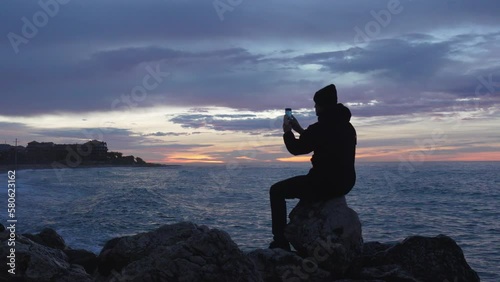 blue hour at sea, man sits on a rock enjoys his time looking at sunset beach, takes photo and leaves photo