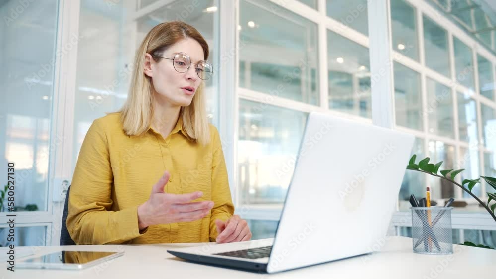 Positive blond mature business woman designer looking at laptop computer screen talking by online distance remote working meeting conference video call conversation using webcam at glass workplace