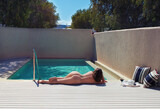Alone beautiful young naked woman lying and sunbathing near the private pool on the wooden terrace with straw hat and glass of wine