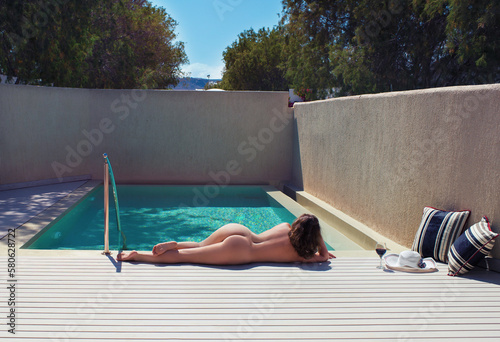 Alone beautiful young naked woman lying and sunbathing near the private pool on the wooden terrace with straw hat and glass of wine