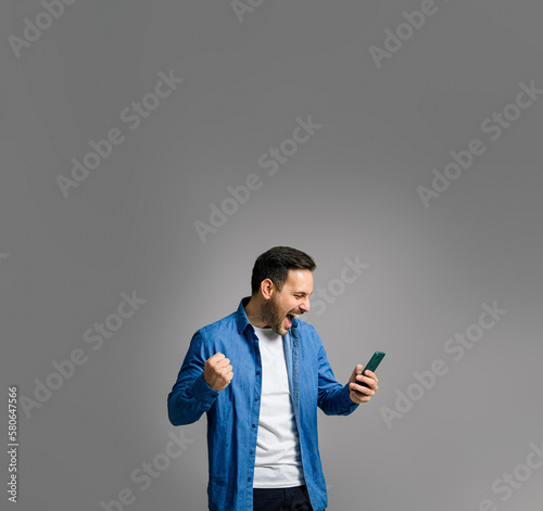 Charming mid adult man in denim shirt shouting ecstatically and pumping fist while reading good news over smart phone isolated on gray background © Moon Safari