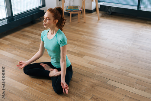 High-angle view of peaceful young woman meditating in lotus position sitting on floor with closed eyes at home enjoying meditation with hands on knee. Concept of healthy lifestyle.