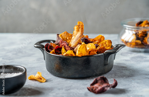 Healthy vegetable chips in a black pot, marine salt, other chips in a glass jar on light surface with grey concrete background