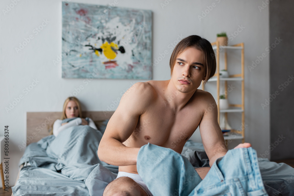 shirtless man holding denim jeans while sitting on bed near blurred woman after one night stand. shirtless man holding denim jeans while sitting on bed near blurred woman after one night stand.