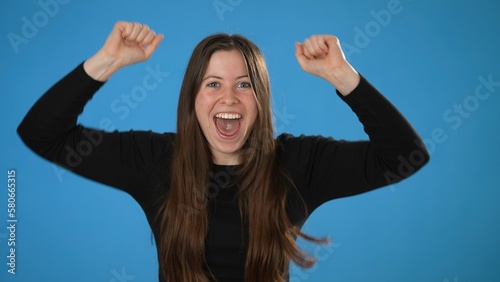 Portrait of excited jubilant overjoyed happy young brunette woman 20s wears black shirt doing winner gesture celebrate clenching fists say yes isolated on plain blue background in studio