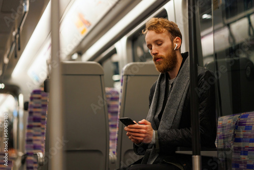 Male subway passenger sits on a train listening to music on headphones and using a smartphone © Andrii 