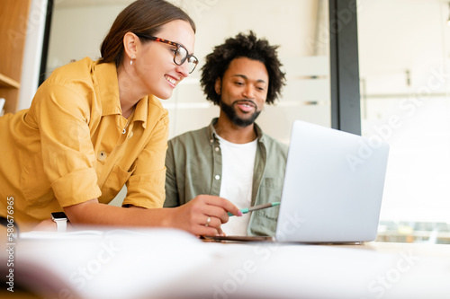 Two colleagues have pleasant conversation sitting side by side in modern open space office in front of laptop, smiling and enjoying friendly working atmosphere, brainstorming together, solving tasks