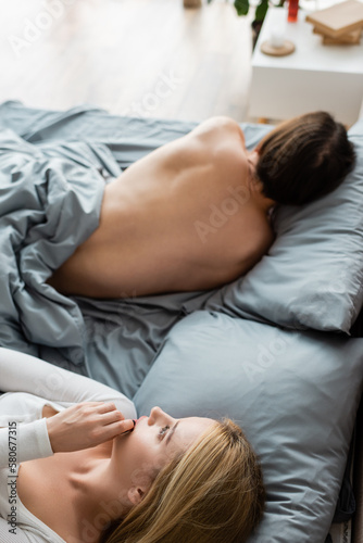 high angle view of embarrassed woman covering mouth while looking at shirtless man in her bed after one night stand.