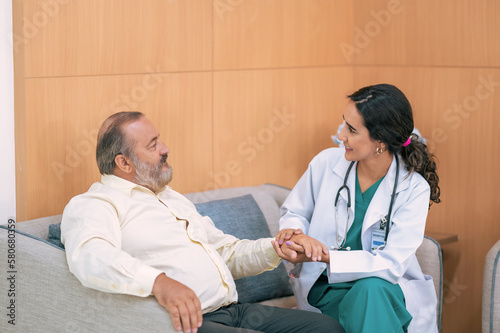 Female doctor in white medical coat and patient discussing something, holding his hand and smiling while sitting on sofa. Medicine and health care concept. Doctor and patient.