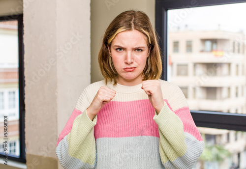 pretty caucasian woman looking confident, angry, strong and aggressive, with fists ready to fight in boxing position. home interior concept