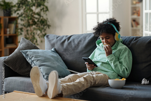 Cute youngster in pants and sweatshirt sitting on sofa while keeping stretched legs on table and watching video in mobile phone © pressmaster