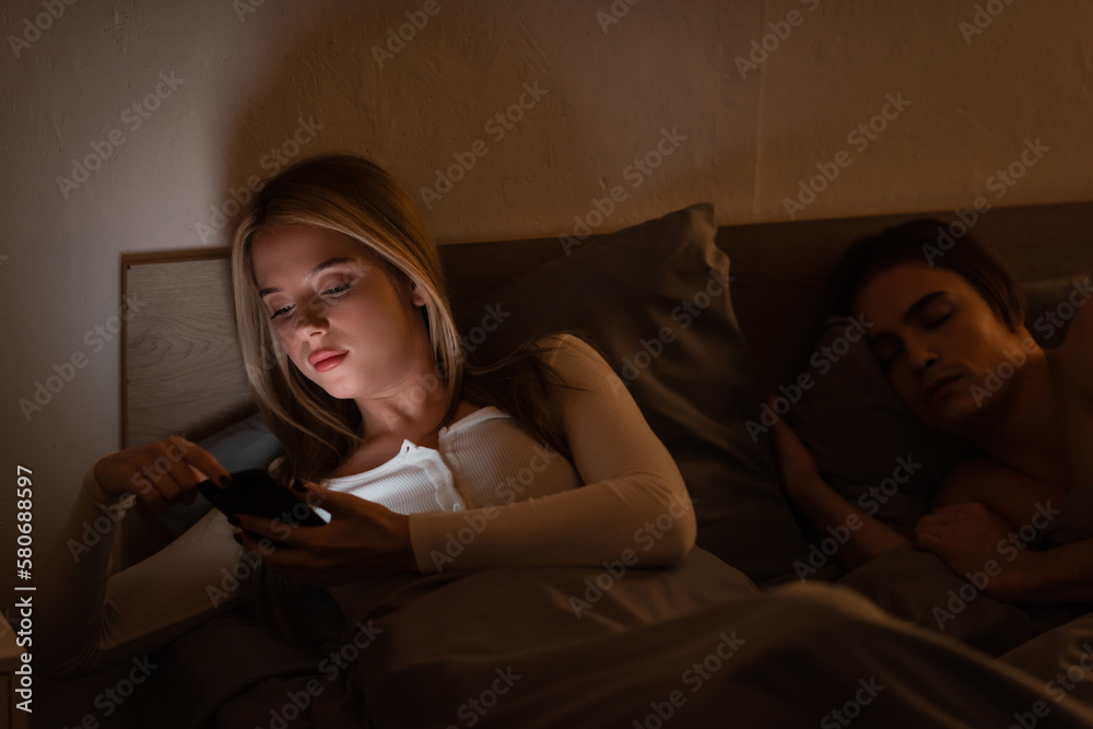 Fototapeta premium unfaithful woman using smartphone next to sleeping boyfriend at night, cheating concept.