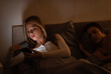 unfaithful woman using smartphone next to sleeping boyfriend at night, cheating concept.
