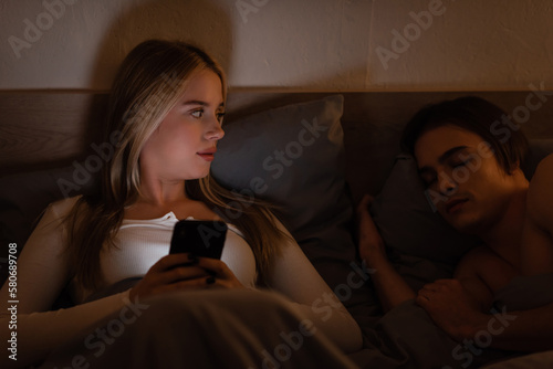 blonde woman using smartphone next to sleeping boyfriend at night, cheating concept.