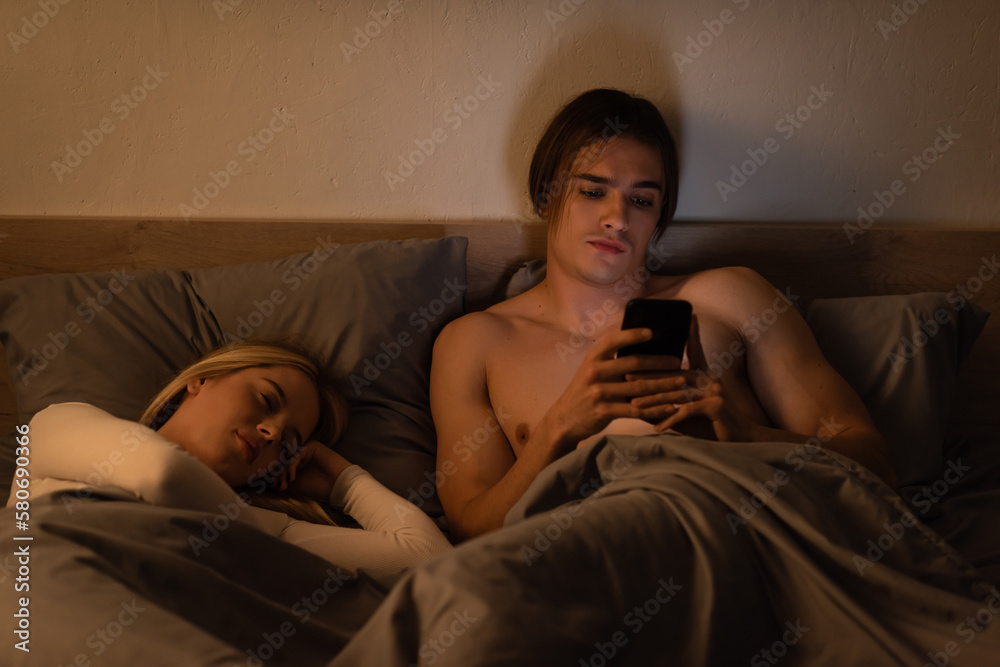 awakened man using mobile phone next to blonde woman sleeping at night, cheating concept.
