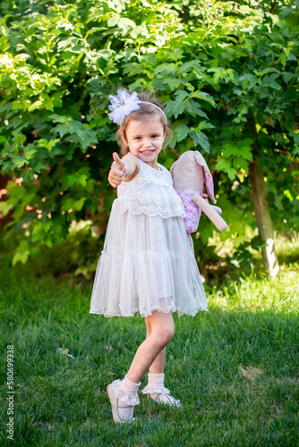 Cute little girl is holding a plush toy, playing in the yard. Beautiful little girl smiles in a smart dress.