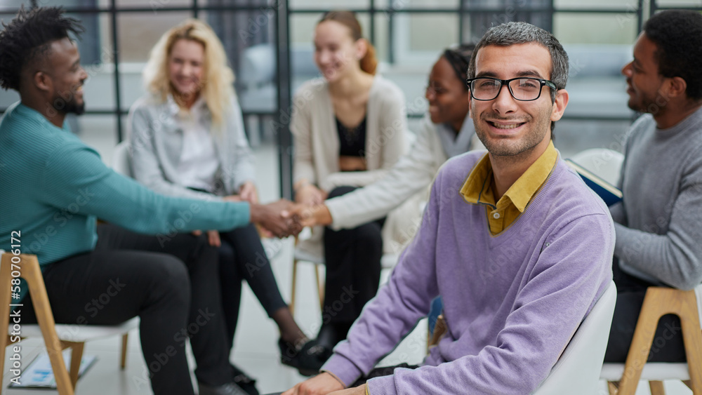 A happy young businessman, in a purple sweater, sits against the background of his colleagues