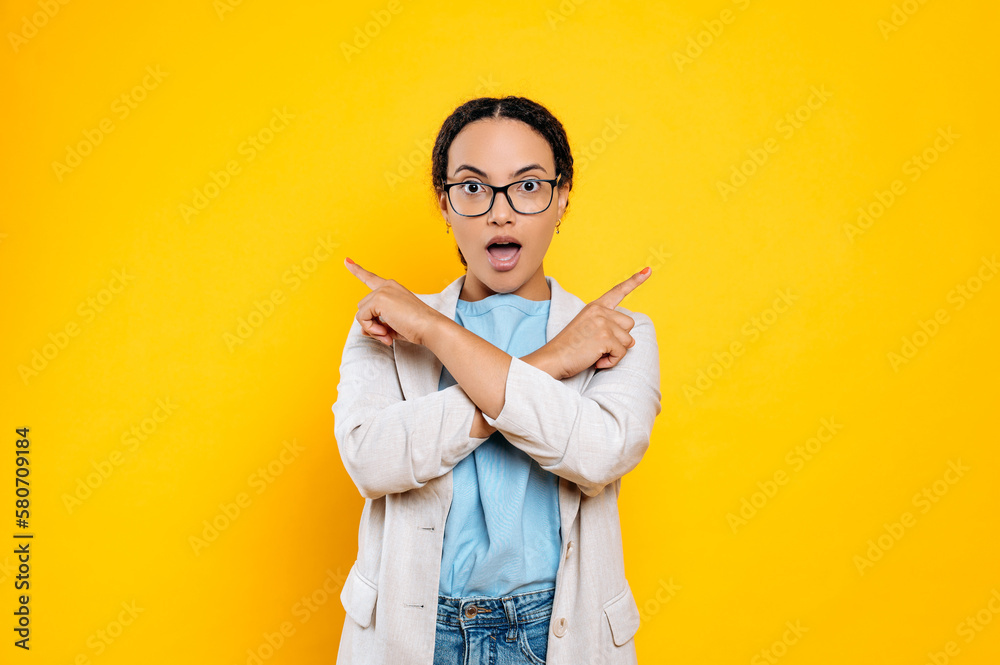 Excited happy mixed race woman with curly hair, wearing stylish clothes, pointing fingers of both hands to the sides at empty space, amazed looks at camera, smile, stands on isolated yellow background Excited happy mixed race woman with curly hair, wearing stylish clothes, pointing fingers of both hands to the sides at empty space, amazed looks at camera, smile, stands on isolated yellow background