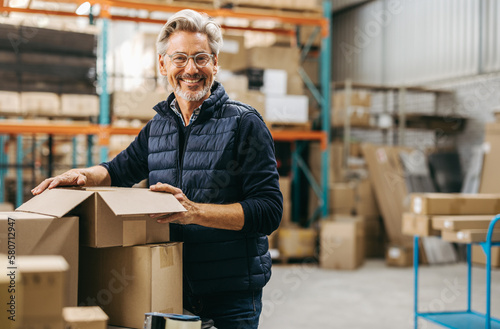 Mature man smiling at the camera while packing cardboard boxes in a warehouse photo