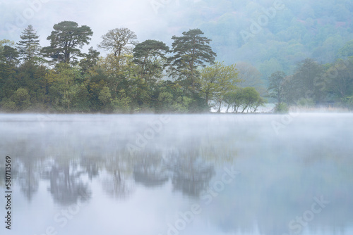 Peaceful morning at lake with mist rising from water and calm reflections of trees. Rydal Water, Lake District, UK.