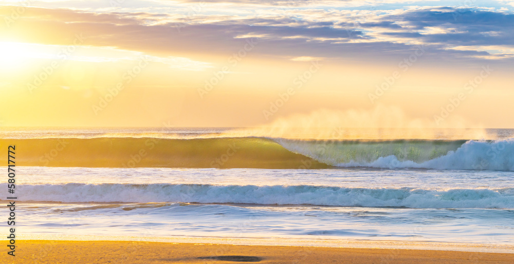 huge beautiful wave is breaking at the coastline while a breeze blows the spit water out of the sea at a wonderful sunset with orange sky