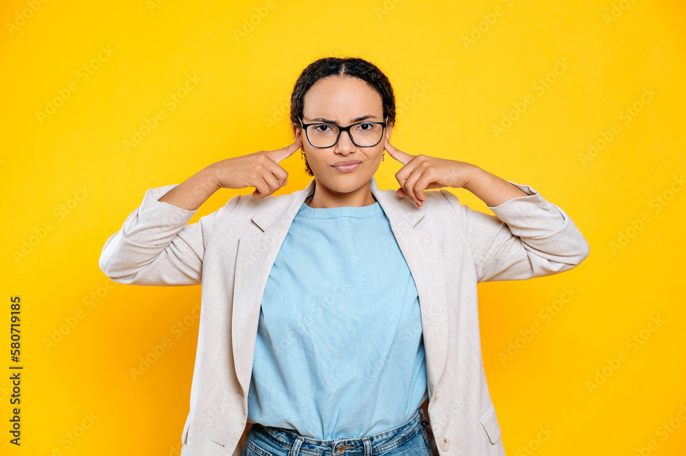 Displeased upset mixed race brunette business woman with glasses, company worker, covering ears with fingers, sadly looks at camera, stand on isolated yellow background Displeased upset mixed race brunette business woman with glasses, company worker, covering ears with fingers, sadly looks at camera, stand on isolated yellow background