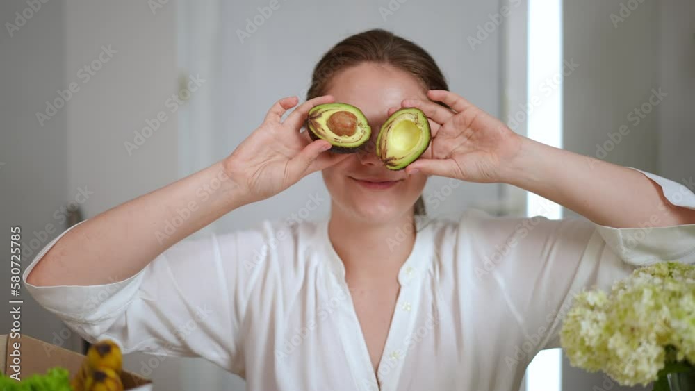 Front view joyful young beautiful woman closing eyes with avocado halves smiling and looking at camera. Cheerful Caucasian housewife having fun posing cooking at home indoors Front view joyful young beautiful woman closing eyes with avocado halves smiling and looking at camera. Cheerful Caucasian housewife having fun posing cooking at home indoors